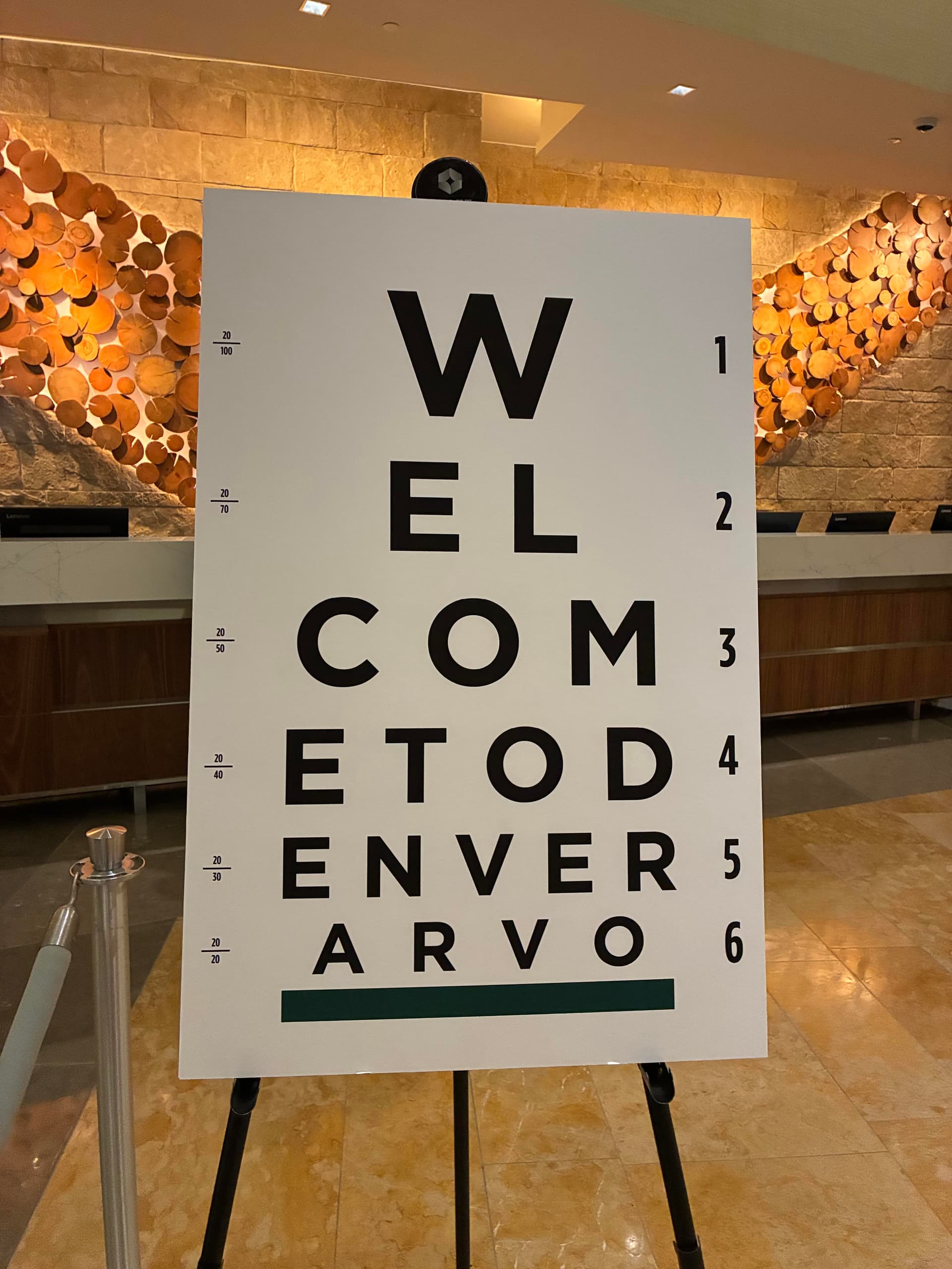 Welcome sign resembling an eye chart, reading "WELCOME TO DENVER ARVO", in a hotel lobby with wood and stone decor.