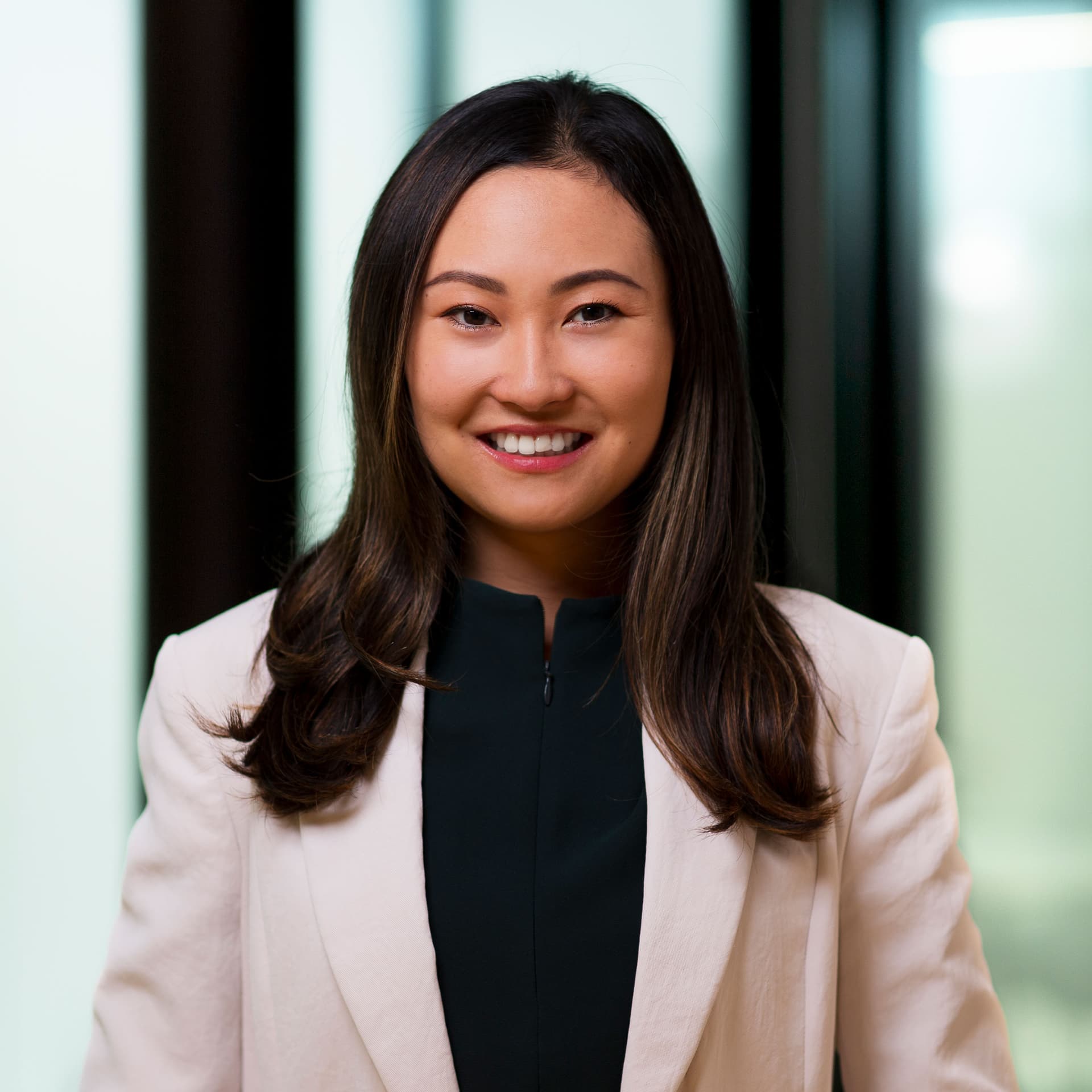 A smiling Asian woman with long dark hair, wearing a dark green top and a light beige blazer