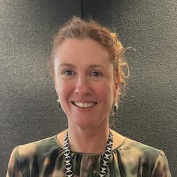 Smiling woman with curly red hair, wearing a patterned top and a dark blue and silver necklace, against a dark background