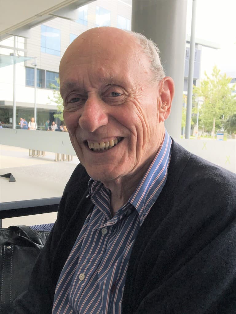 A smiling older man with a bald head and blue striped shirt wearing a dark cardigan, sitting outdoors