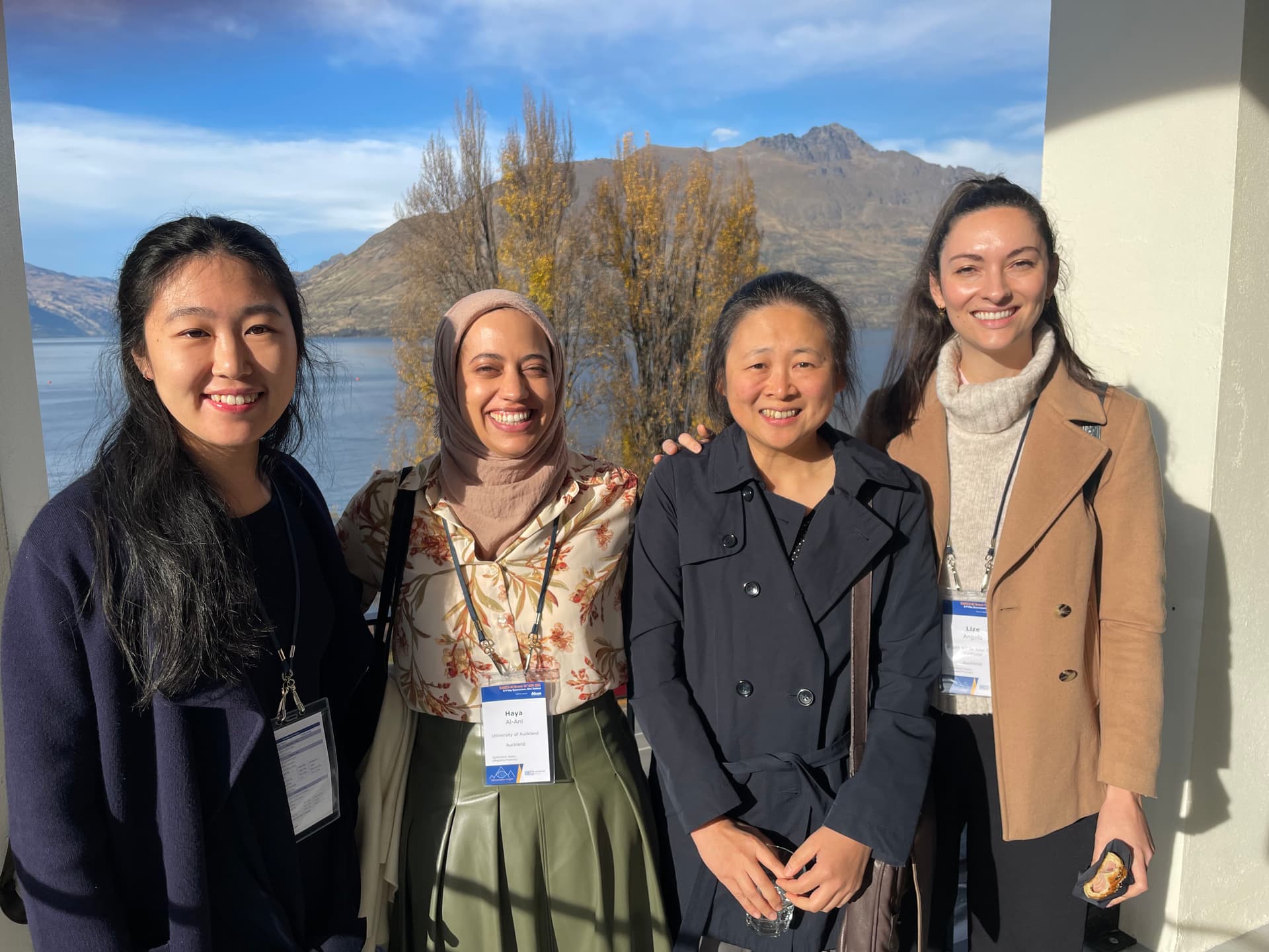 Four women, smiling, stand on a balcony with a lake and mountains in the background.