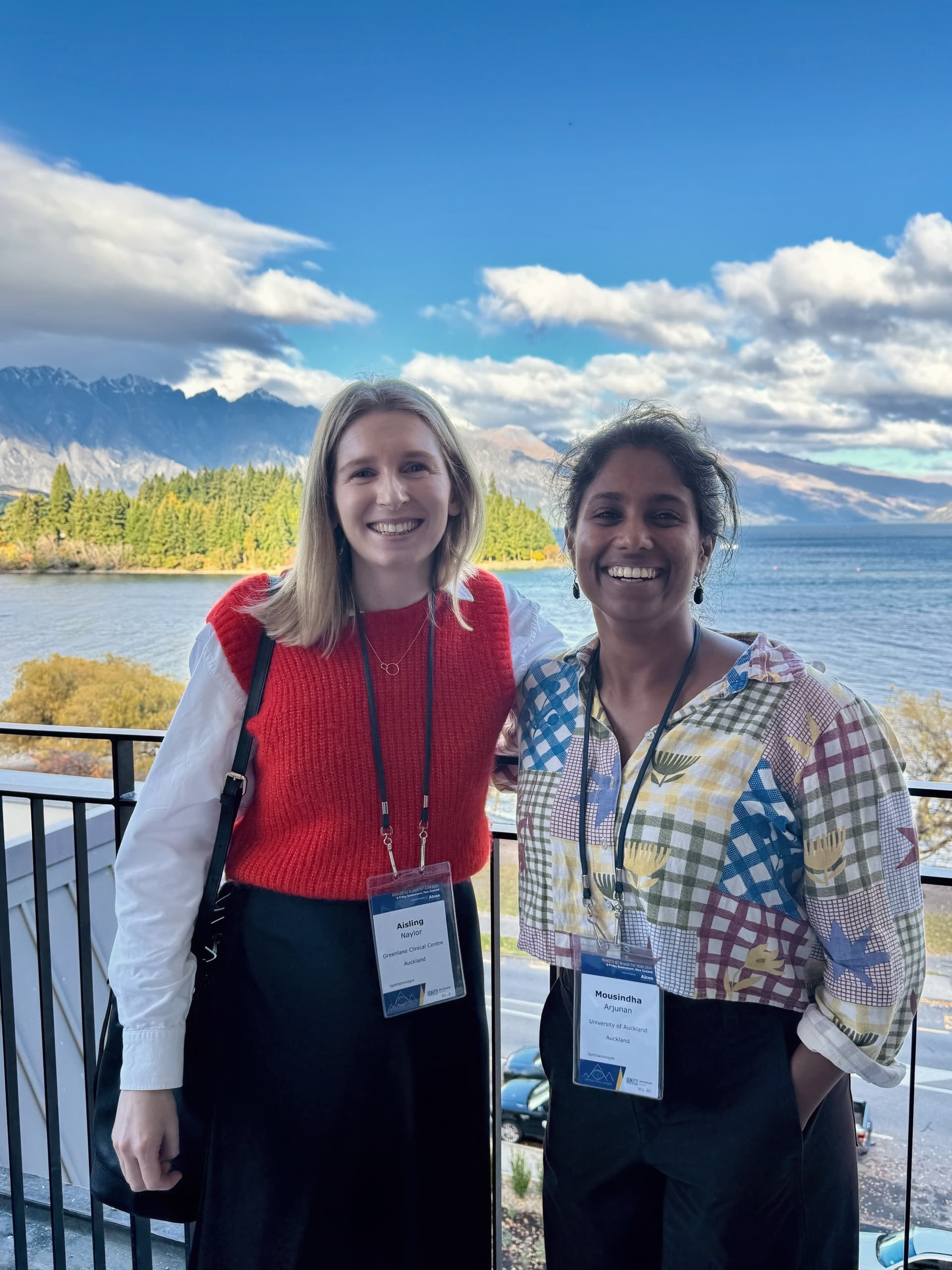 Two women, Aisling Naylor and Mousindha Arjunan, smile at the camera on a balcony overlooking a lake and mountains.