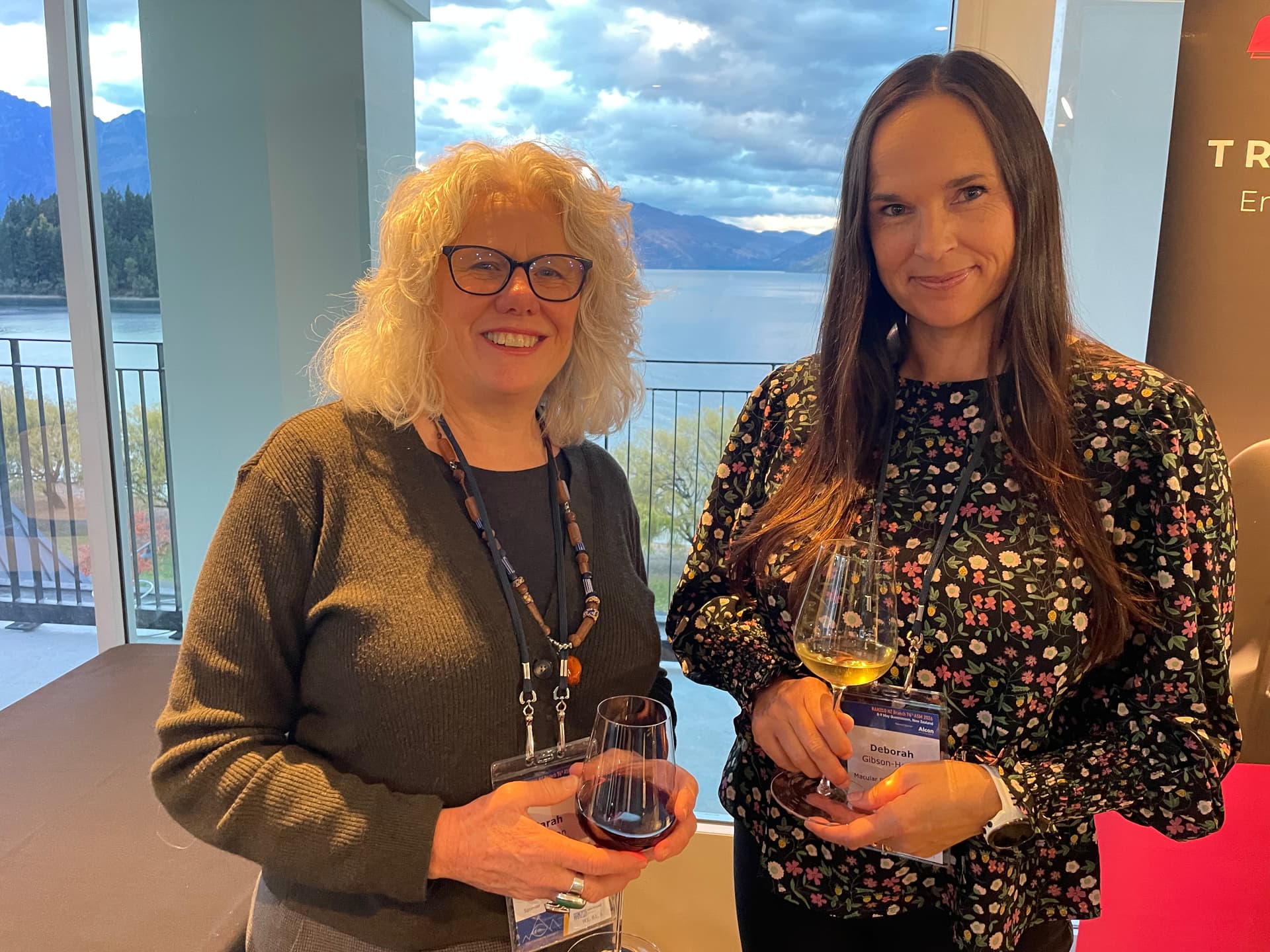 Two women holding wine glasses smile at the camera with a lake and mountains in the background.