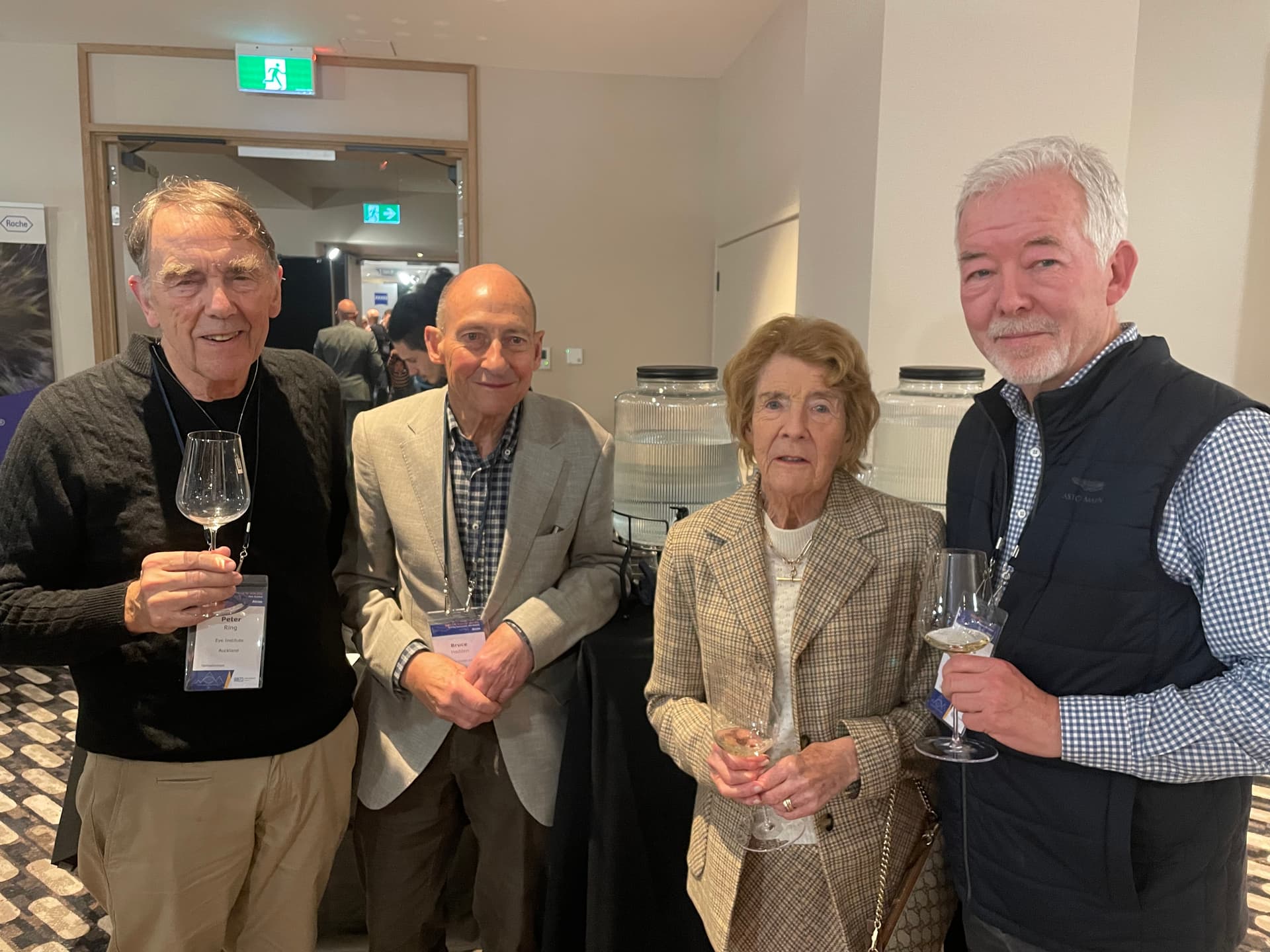 Four senior people, two men and two women, stand smiling with drinks at a conference.