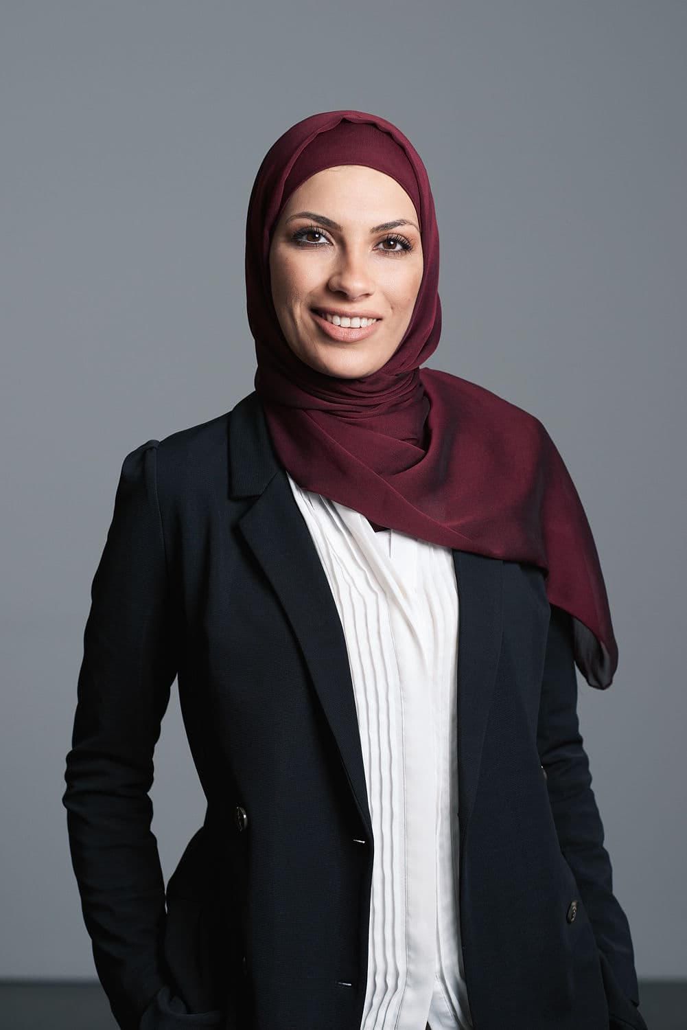 Smiling woman in a dark blue blazer, white pleated shirt, and a maroon hijab, looking directly at the camera