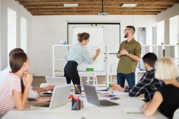 A woman writes on a whiteboard while a man watches, and others work on laptops at a table.