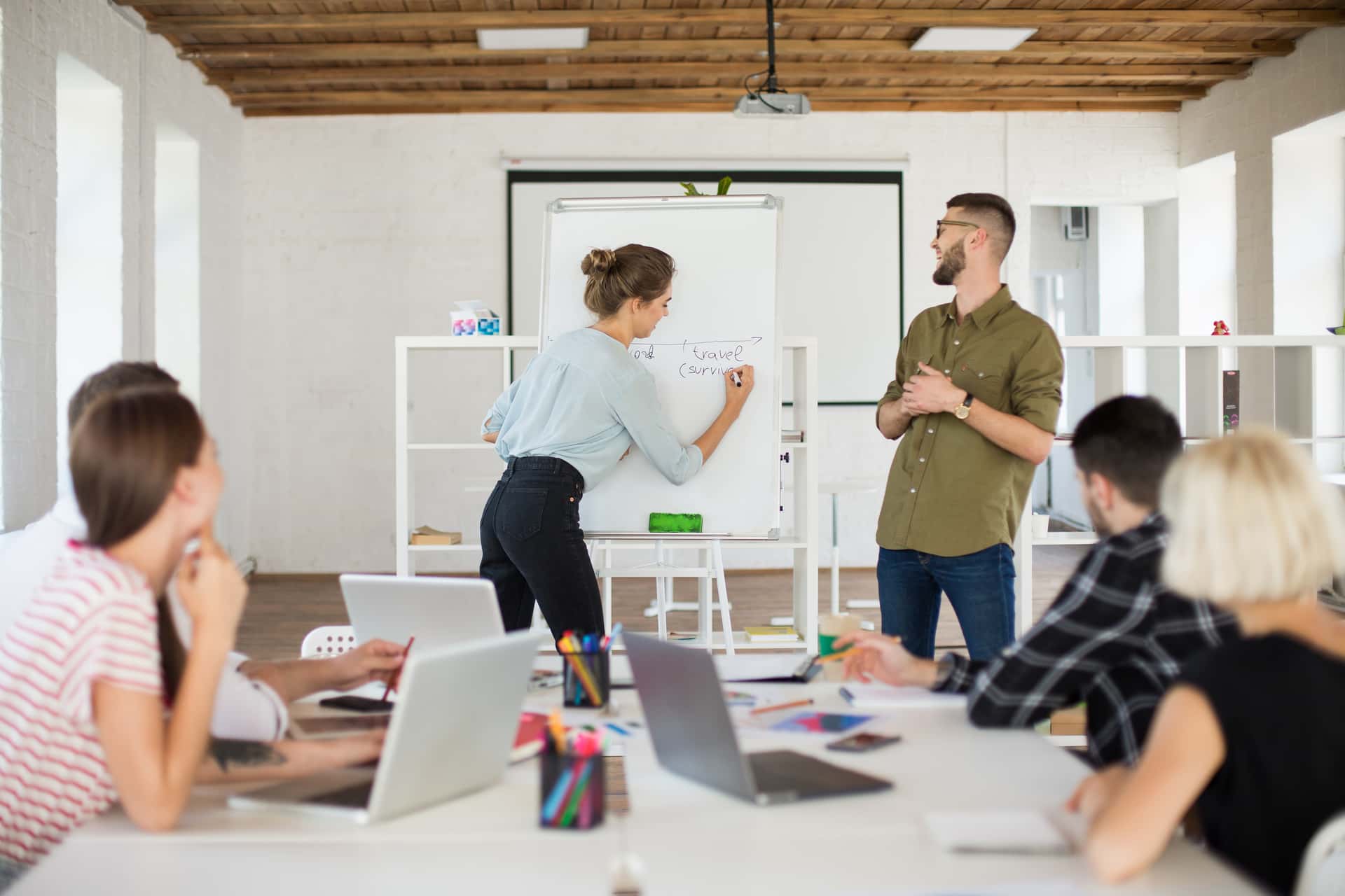 A woman writes on a whiteboard while a man watches, and others work on laptops at a table.